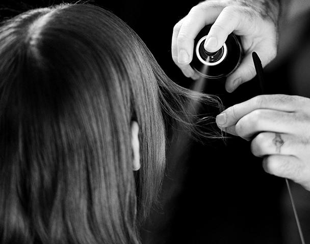 The back of a woman's head with a strand of dark hair held out as hairspray is applied