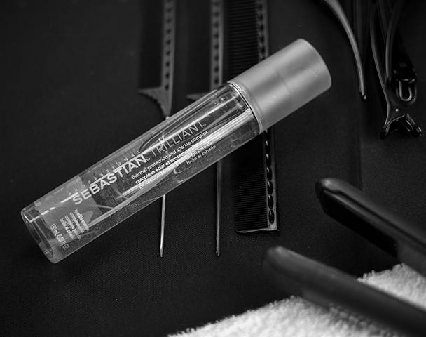 Black and white photo of a slim bottle of Trilliant heat protection spray, lying on top of 3 combs