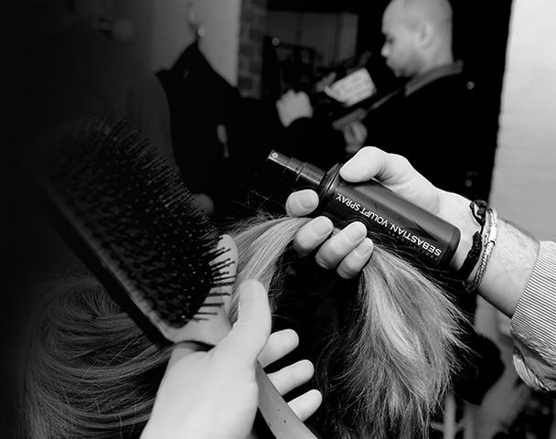 The back of a woman's head, as hands hold a section of her blonde hair and a hairbrush